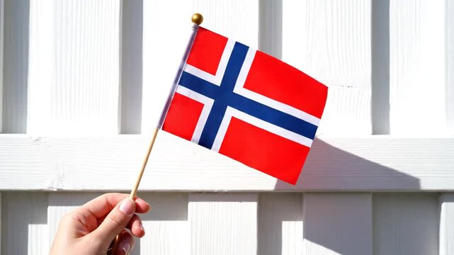 A hand holding a small Norwegian flag casts a shadow against a white wooden grid background, with multiple similar images arranged in a pattern.