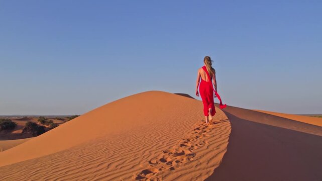 Aerial view of a woman in a long red dress walking on the crest of a sand dune, with the bright red contrasting against the orange sand, Carlsbad, New Mexico, United States.