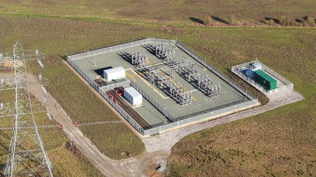 Aerial view of a grey substation with silver electrical equipment contrasting against the brown grass, bordered by a tall pylon, Luton, United Kingdom.