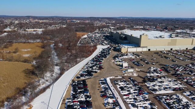 Aerial view of a large parking lot filled with cars next to a mall, contrasting with a snowy landscape under a clear blue sky, Framingham, Massachusetts, United States.