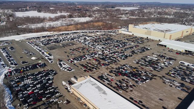 Aerial view of a vast parking lot filled with cars next to large buildings, contrasting the asphalt with surrounding snow, Framingham, Massachusetts, United States.