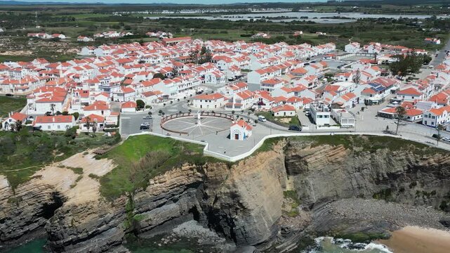 Aerial view of the village, with its white buildings and red roofs, nestled against the rugged cliffs overlooking the sea, Zambujeira do Mar, Beja District, Portugal.