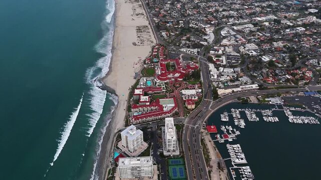 Aerial view of the iconic red-roofed Hotel del Coronado, set against the blue ocean and sandy beach, contrasting with the marina, Coronado, California, United States.