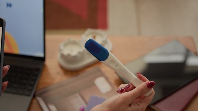 Close up shot of woman holding pregnancy test while checking menstrual cycle application on smartphone in front of laptop on wooden table at home