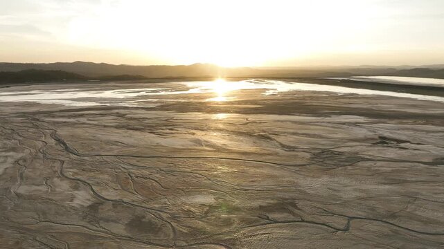 Aerial view of a cracked, dry riverbed reflecting the sun's golden light, contrasting with the distant water and mountains, Minas de Riotinto, Andalusia, Spain.