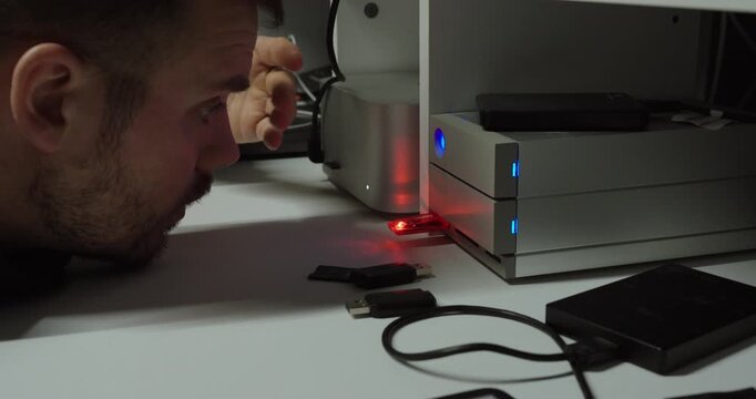 Man closely observing a desktop storage device with blinking red and blue indicator lights, suggesting slow data transfer, system performance issues, or hardware troubleshooting in a home office setup