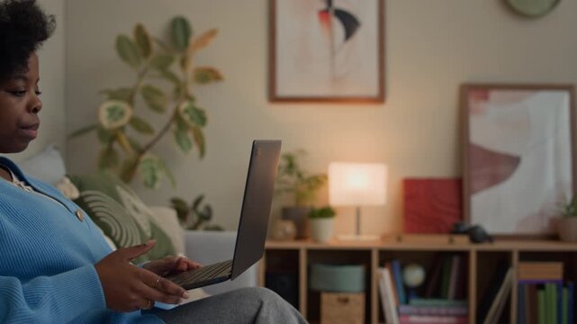 Tracking shot of young Black woman monitoring health indicators in application on wireless laptop while sitting on sofa in living room