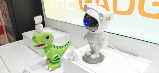 Fototapeta premium 6 December 2025, Abu Dhabi, UAE: Green T Rex toy standing next to a futuristic astronaut galaxy projector on a white retail display, capturing modern gadgets