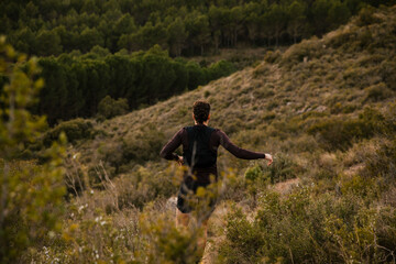 Obraz premium Rear view of a trail runner moving along a rugged mountain trail surrounded by wild nature and forested hills. Outdoor adventure sport and endurance lifestyle concept.