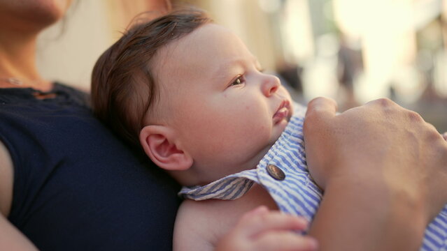 Close-up of baby lying on mother&rsquo;s lap outdoors, looking directly into camera with curious expression, relaxed in arms during family outing in urban setting