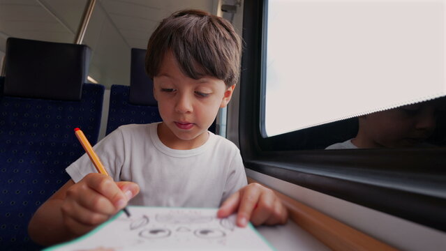 Young boy drawing with pencil on paper inside train, looking down with focus, window reflection visible, blue seat, travel, creative activity during journey