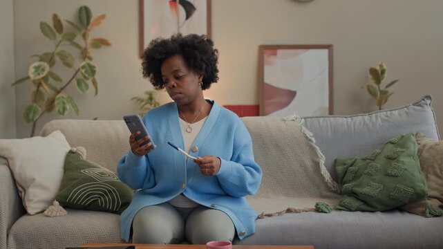 Medium long shot of young Black woman getting result of pregnancy test and checking smartphone while sitting on cozy sofa in modern living room with abstract paintings in background