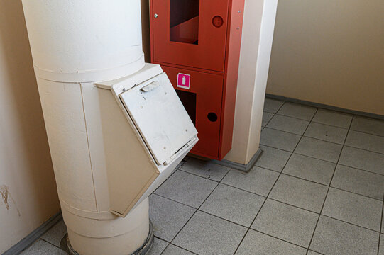 A white cylindrical waste chute with a hinged metal flap door mounted on a pillar, adjacent to a red fire safety cabinet with a fire extinguisher sign, in a tiled corridor