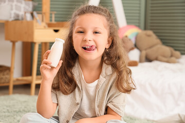 Fototapeta premium Cute little girl with bottle of yogurt showing tongue in room