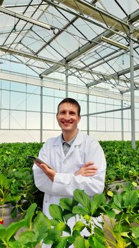 Smiling researcher in a white lab coat stands in a greenhouse among leafy crops, holding a smartphone, showcasing modern plant science and controlled-environment agriculture.