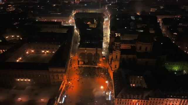 Night aerial drone footage showing fireworks above Munich historic center, including Odeonsplatz and Ludwigstrasse. City lights, traffic lines and winter urban environment seen from a high angle.