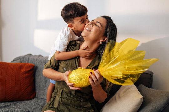 Brazilian mother surprising young son with handmade Easter egg gift on living room sofa, celebrating family affection, childhood joy and traditional holiday moments at home.