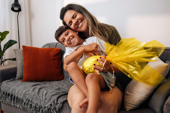 Brazilian mother surprising young son with handmade Easter egg gift on living room sofa, celebrating family affection, childhood joy and traditional holiday moments at home.