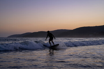 Silhouette of a Surfer Riding Ocean Waves at Sunset with Dramatic Sky and Distant Hills in the Background