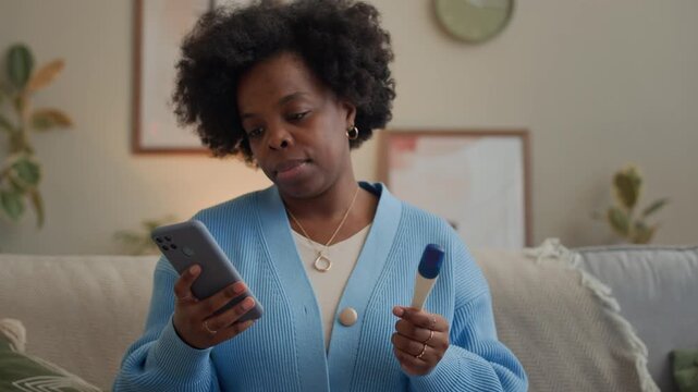 Tilt up shot of young adult African American woman in blue cardigan holding pregnancy test and using mobile phone while sitting on sofa in living room