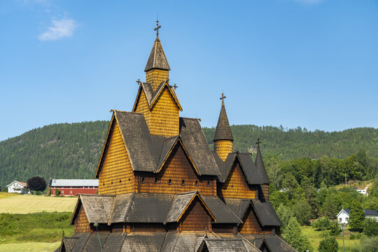 View of a golden-brown wooden Heddal stave church rises majestically against a clear blue sky, framed by rolling green hills, Heddal, Telemark, Norway.