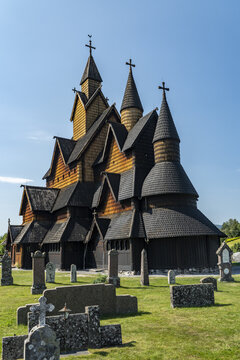View of the Heddal Stave Church, a towering wooden structure with intricate carvings, stands amidst weathered gravestones under a clear blue sky, Heddal, Telemark, Norway.