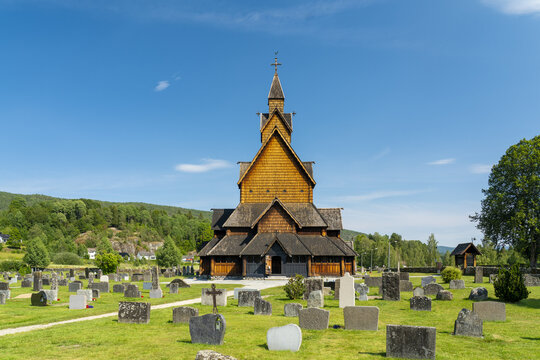 View of a majestic wooden Heddal stave church rises against a clear blue sky, surrounded by a historic graveyard with weathered stones, Heddal, Telemark, Norway.