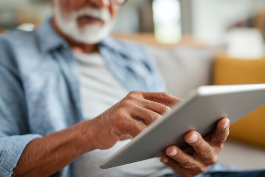 Senior man uses tablet with focused expression. He sits comfortably on a couch, dressed casually. Soft lighting highlights his hands and device. The tablet is held securely