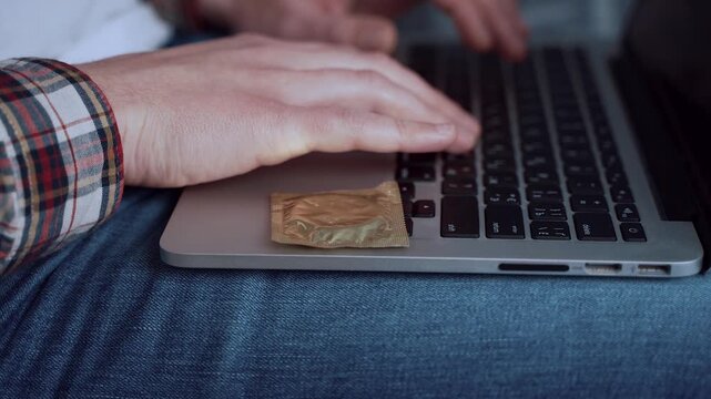 Close up of man typing on computer with a condom on the edge. Importance of protection in one-night stands and casual sexual relationships. Safe health practices for anonymous online meetings.