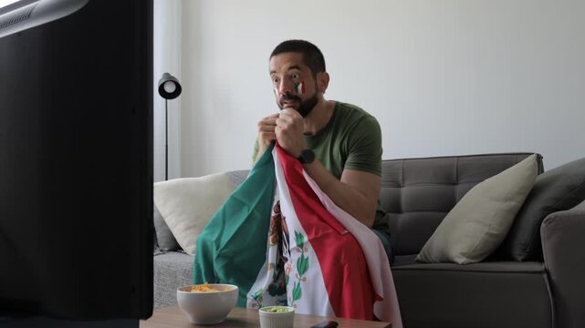 Mexican football fan celebrating at home while holding national flag and watching a match on TV. Adult man cheering on the sofa with snacks and drinks in the living room.