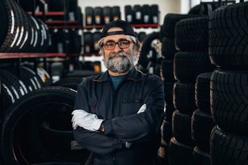 Fototapeta premium Male auto technician with glasses and gray beard stands confidently among stacks of tires in a tire shop, wearing a dark coverall and gloves