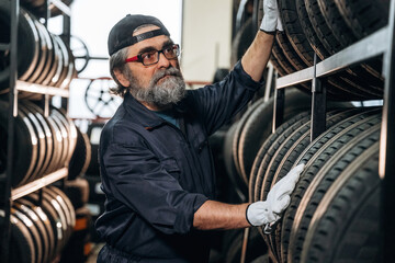 Caucasian male mechanic with glasses adjusts tire on rack inside a tire shop, stacks of tires and workshop tools visible in the background © standret