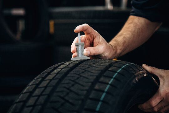 Hand of a technician measuring tire tread depth with a gauge on a black tire in a workshop surrounded by additional tires in the background