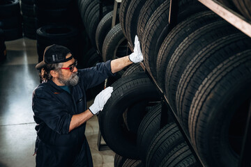 Male tire technician wearing gloves inspects tires on a display rack inside a tire shop, with multiple tires stacked in the background © standret