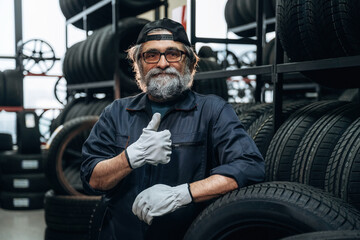 Fototapeta premium Elderly Caucasian male tire technician wearing gloves poses with thumbs up in a tire shop surrounded by stacks of tires and wheels in the background