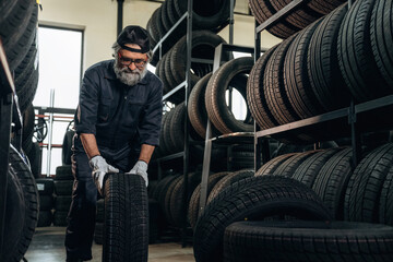 Senior male mechanic with gray beard lifts a tire in a tire storage area, surrounded by shelves filled with various tires and bright natural light from windows