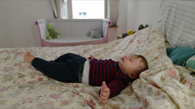 Baby lying on bed complaining with arms outstretched, seeking attention and comfort, early signs of fussiness in cozy family bedroom with soft bedding and natural light