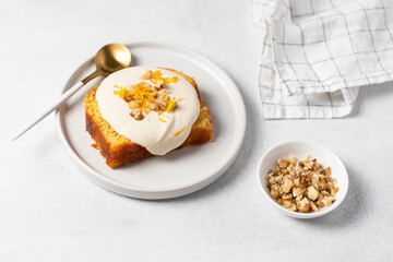 Orange Zest Cream-Topped Cake Slice with Nuts. A golden sponge cake slice topped with a smooth white frosting, garnished with bright orange zest and chopped nuts. Plated on a white round dish.