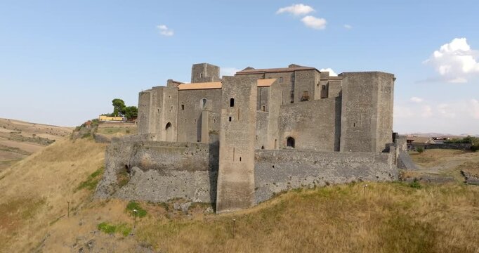 Panorama of the Castle of Melfi in Basilicata, Italy. It is a Norman Fortress, perched atop a grassy hill. The medieval fort, with its high walls and square towers, stands against a clear blue sky.