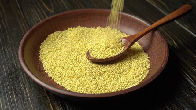 Yellow millet grains being poured from a wooden spoon into a brown bowl on a dark wooden surface, showcasing the pouring action and grain texture