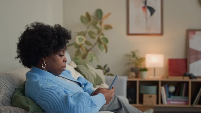 Zoom out shot of relaxed African American woman monitoring menstrual period in calendar on smartphone while resting on sofa at home