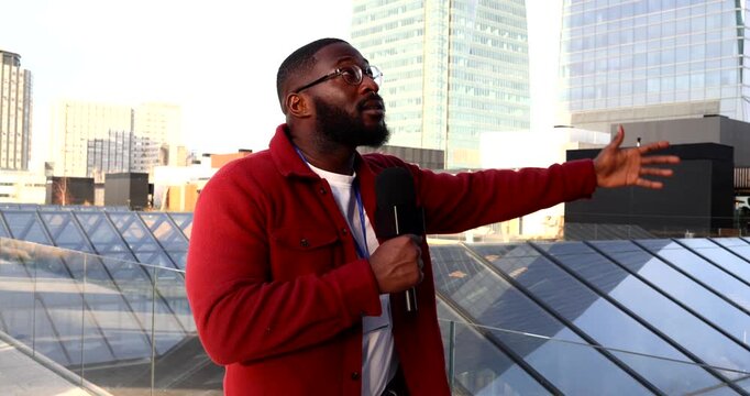 Professional african american male correspondent speaking into a microphone and gesturing while reporting news from a city rooftop with modern skyscrapers in the background on a sunny day