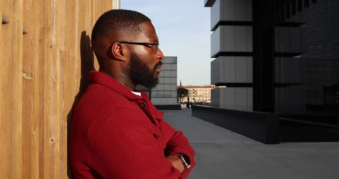 Thoughtful black man with a beard and glasses leaning against a wooden wall, he uncrosses his arms and looks at his smartwatch while waiting in a modern urban architectural setting