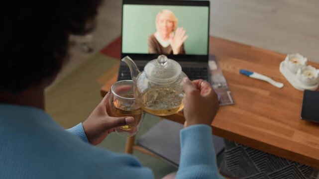 Tracking shot of young African American woman pouring herbal tea into cup while chatting with female Caucasian friend via video call on wireless laptop at home