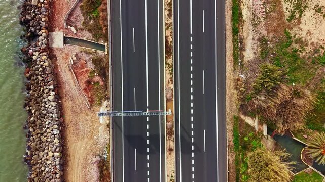 A highway runs parallel to a body of water. Cars travel on the road. On one side, there are rocks and on the other, there are trees. The scene shows a clear day.