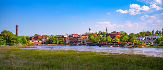 Panoramic view of historic Exeter, New Hampshire, seen across the Squamscott River with brick buildings, church steeples, and summer greenery under a blue sky. © Nick Fox