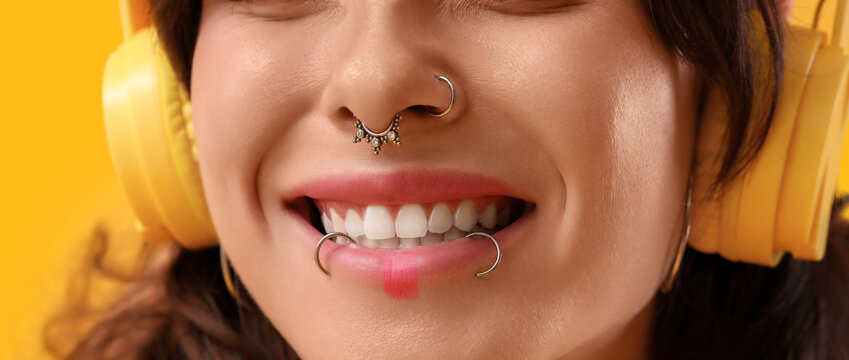Young woman with face piercing listening to music on yellow background, closeup