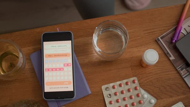 Top down shot of hand of unrecognizable Black woman taking smartphone with application for menstrual cycle tracking from wooden table with pills and cups