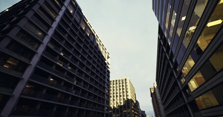 Naklejka premium upward view of clustered highrises under dramatic sky, textured facades catching late light, vertical geometry and repetitive windows creating bold