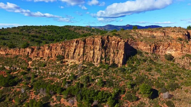 Cliffs rise above a patchy landscape filled with green trees. A bright blue sky with clouds stretches overhead. This scene shows natural beauty in a rural area.
